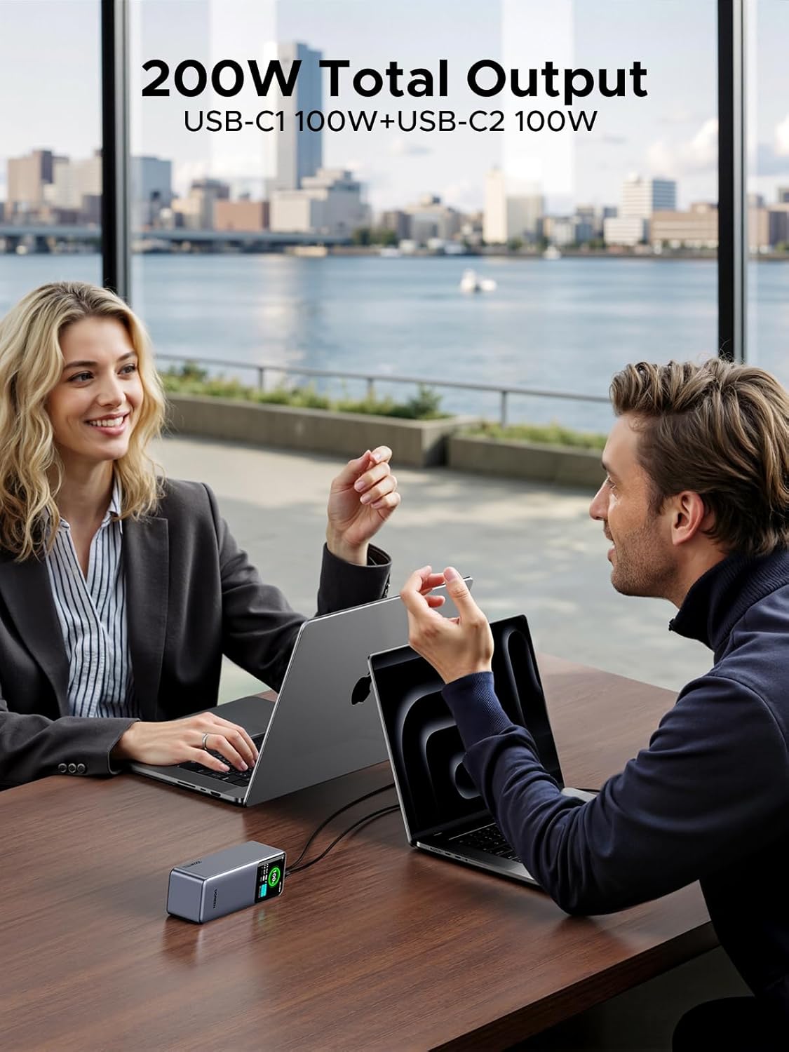 Two people working on laptops with a laptop power bank on a desk by a window with a cityscape view.